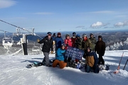A group of 10 veteran skiers, snowboarders and instructors gather atop Mount Ellen. A clear blue sky and views of the surrounding mountains behind the group. Two veterans crouch in front of the rest holding a Vermont Adaptive Ski and Sports banner between them