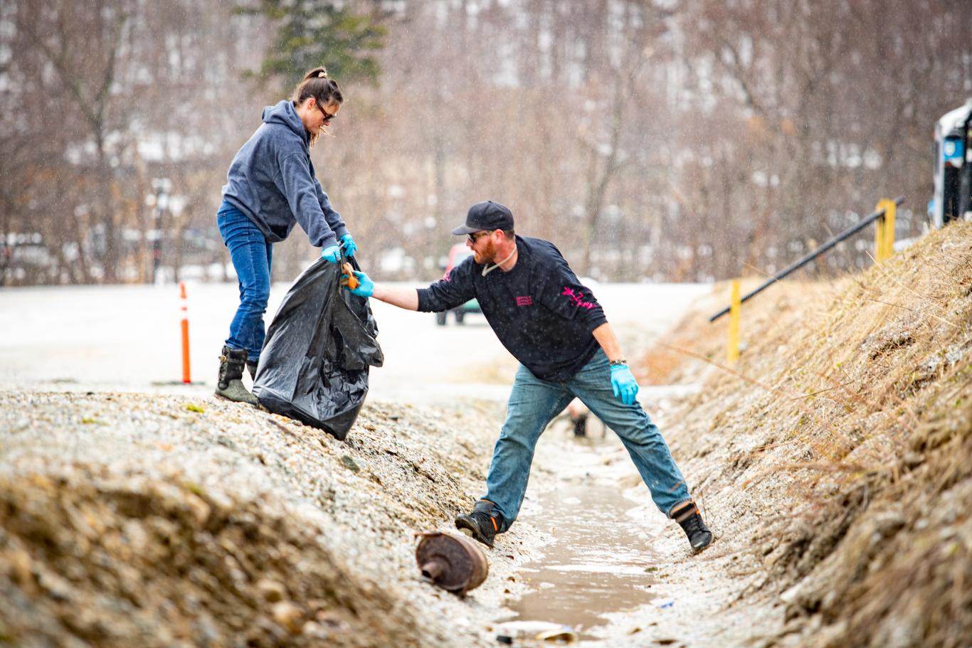 A man standing in a drainage ditch hands trash to a woman extending a trash bag to him