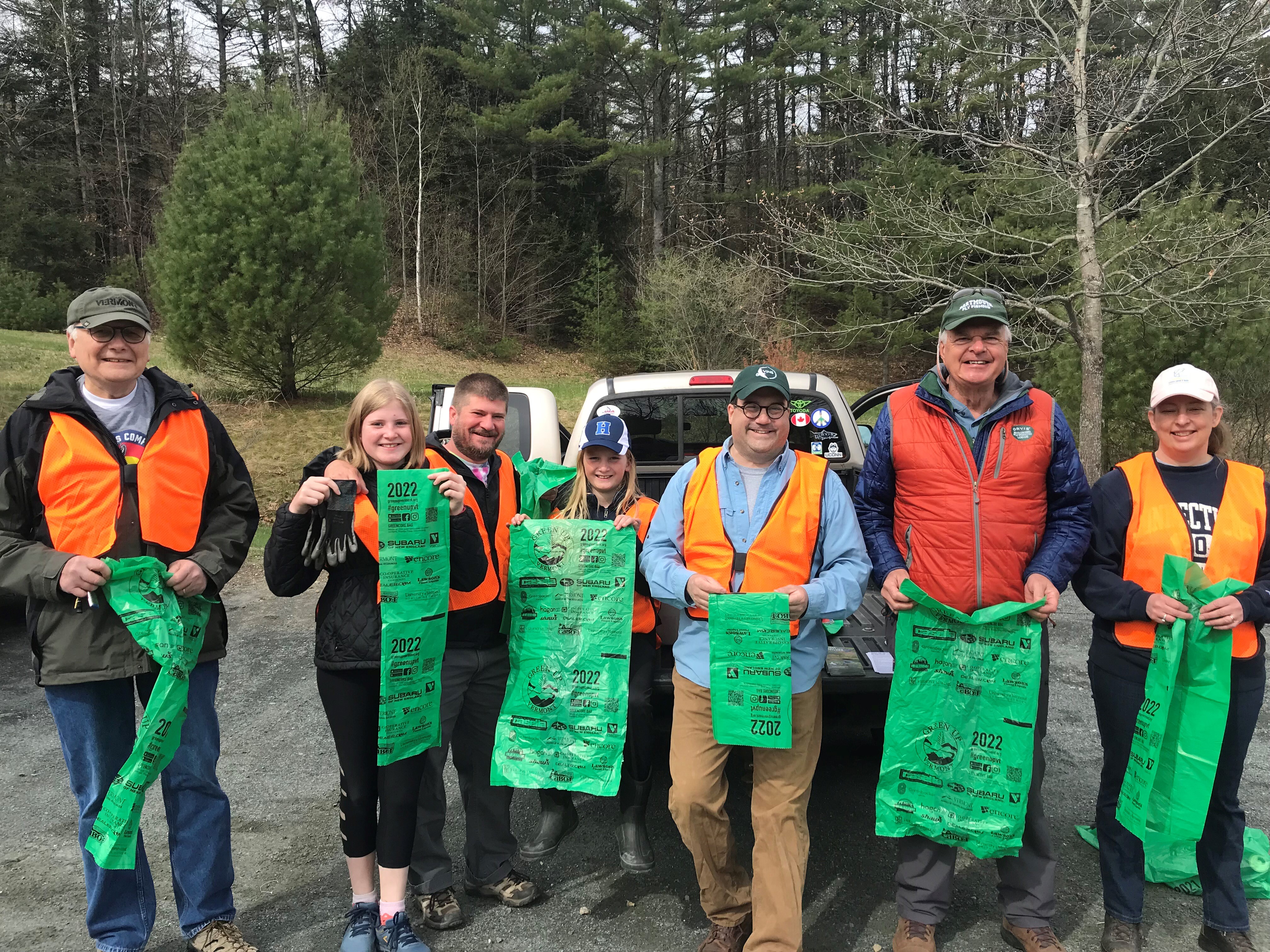 A group of men, women and children wearing reflective vests pose in front of the trash they've cleaned from roadways.